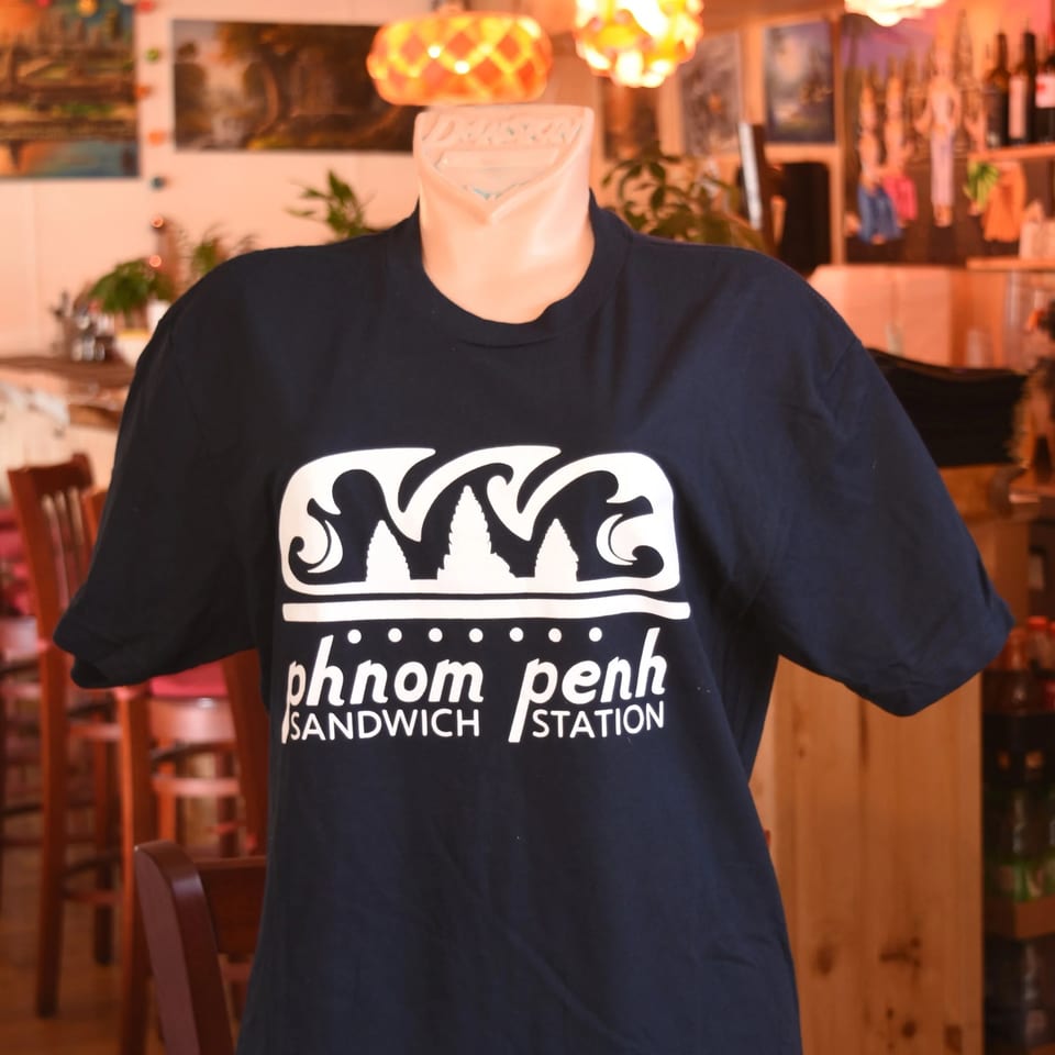 Dark blue Phnom Penh Sandwich Station t-shirt with white logo on a mannequin in a restaurant.