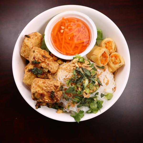 Cambodian noodle bowl with fried tofu, spring rolls, herbs, and a side of dipping sauce.