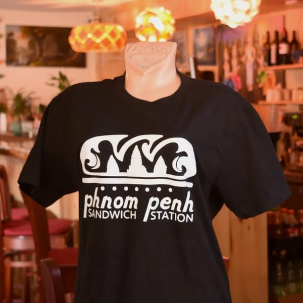 Black t-shirt with "Phnom Penh Sandwich Station" logo on a mannequin in a restaurant.